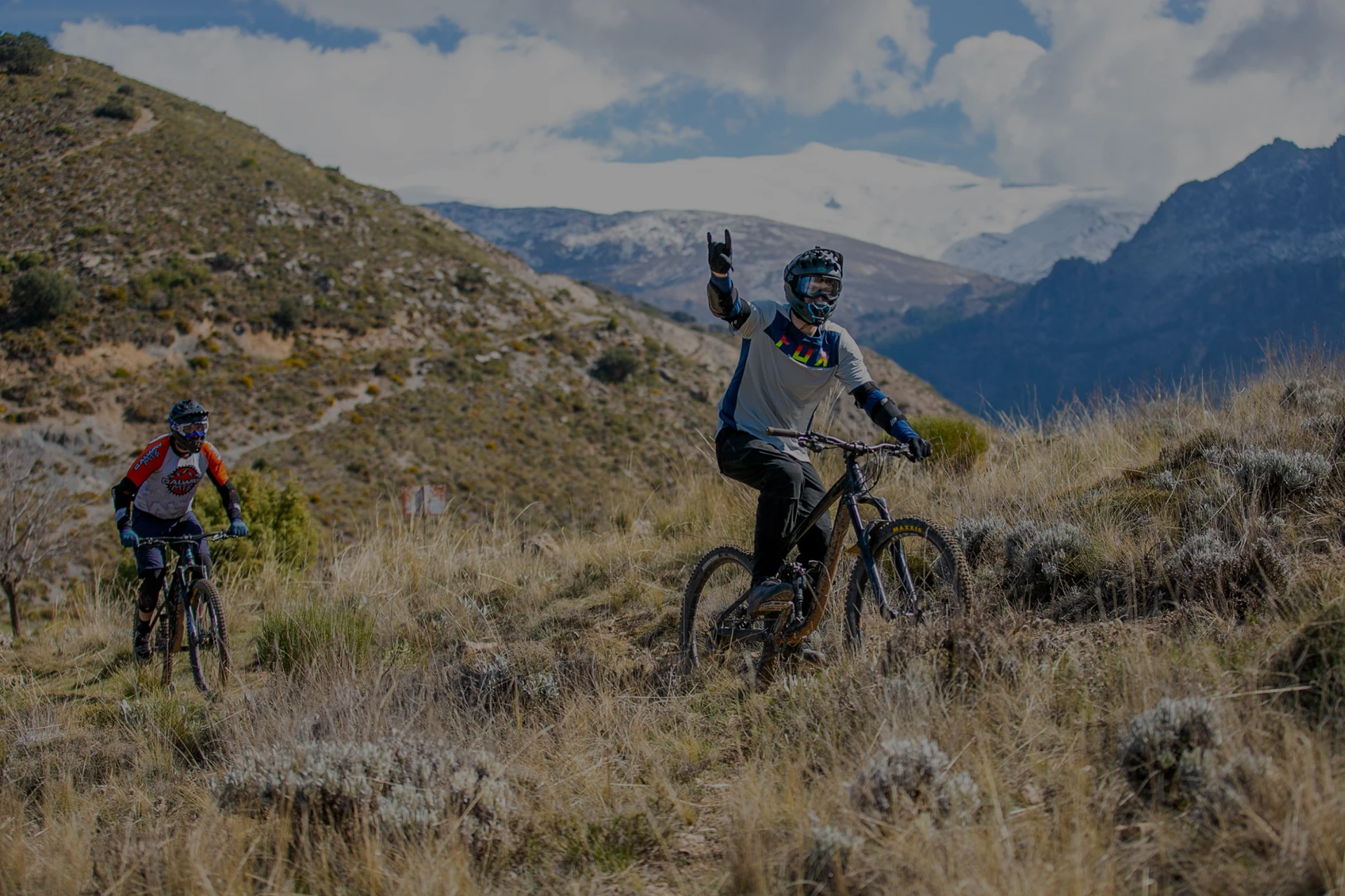 Image of two mountain bikers as they climb the side of a sierra and one of them looks at the camera raising his arm while making horns with his right hand